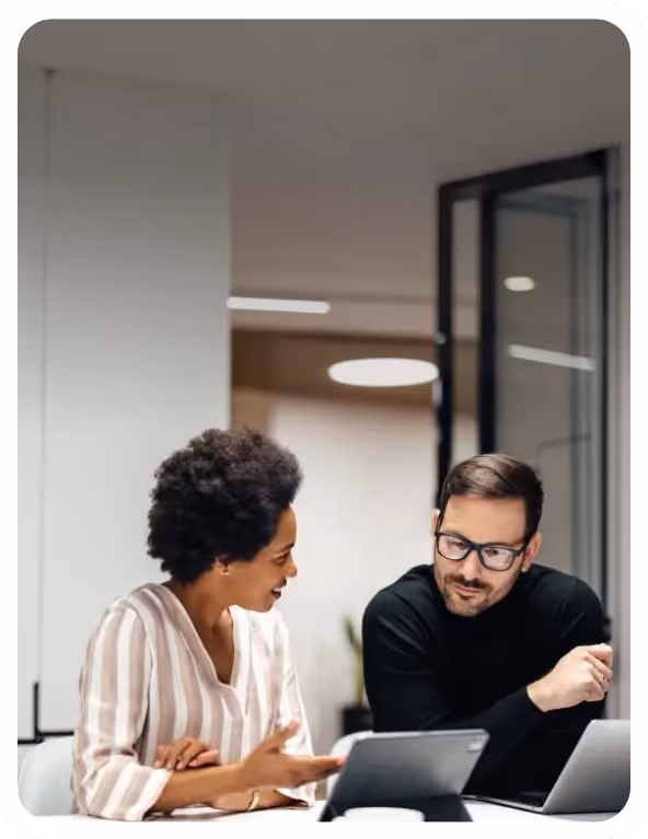 Two colleagues sitting at a desk discussing work while looking at a tablet.
