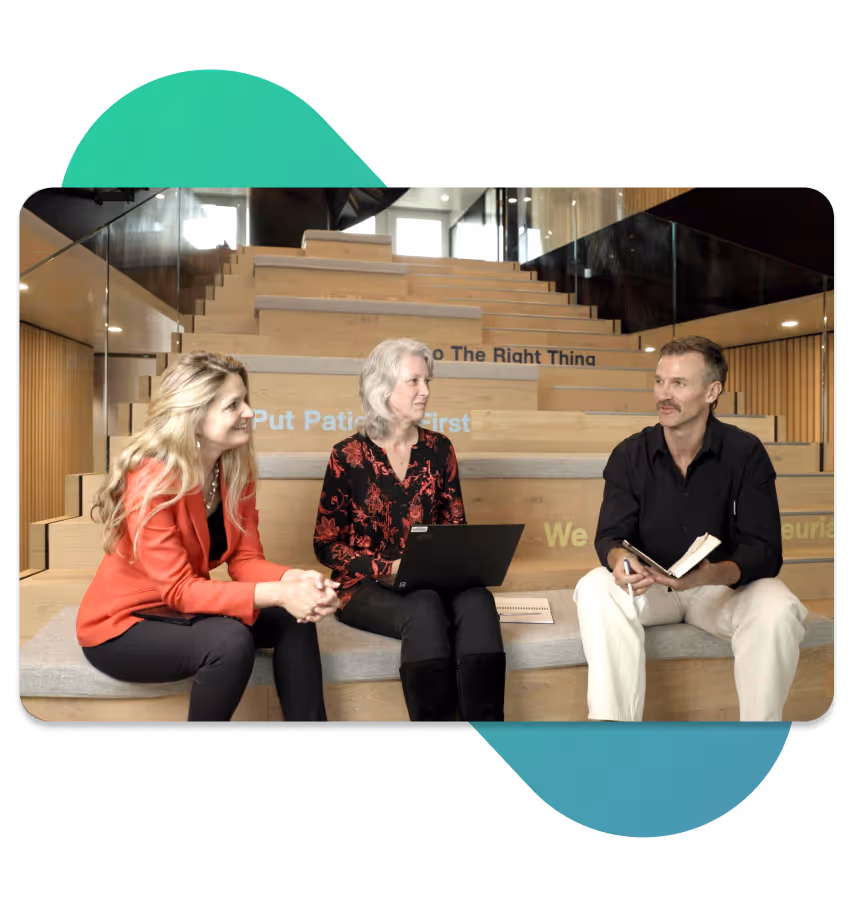 Two women and one man sitting on wooden steps in a modern office, engaged in discussion with a laptop and notebook.