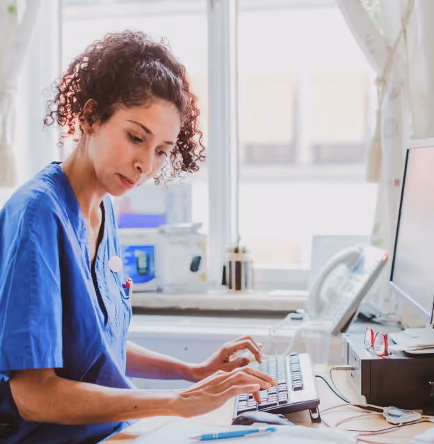 Healthcare professional in blue scrubs working on a computer at a medical office desk.