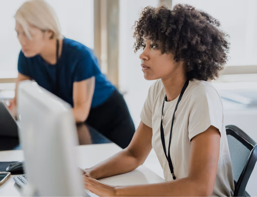 **Alt text:**
Two professionals working at a desk in an office. The person in the foreground is focused on a computer screen, while the person in the background leans forward, also engaged in work.