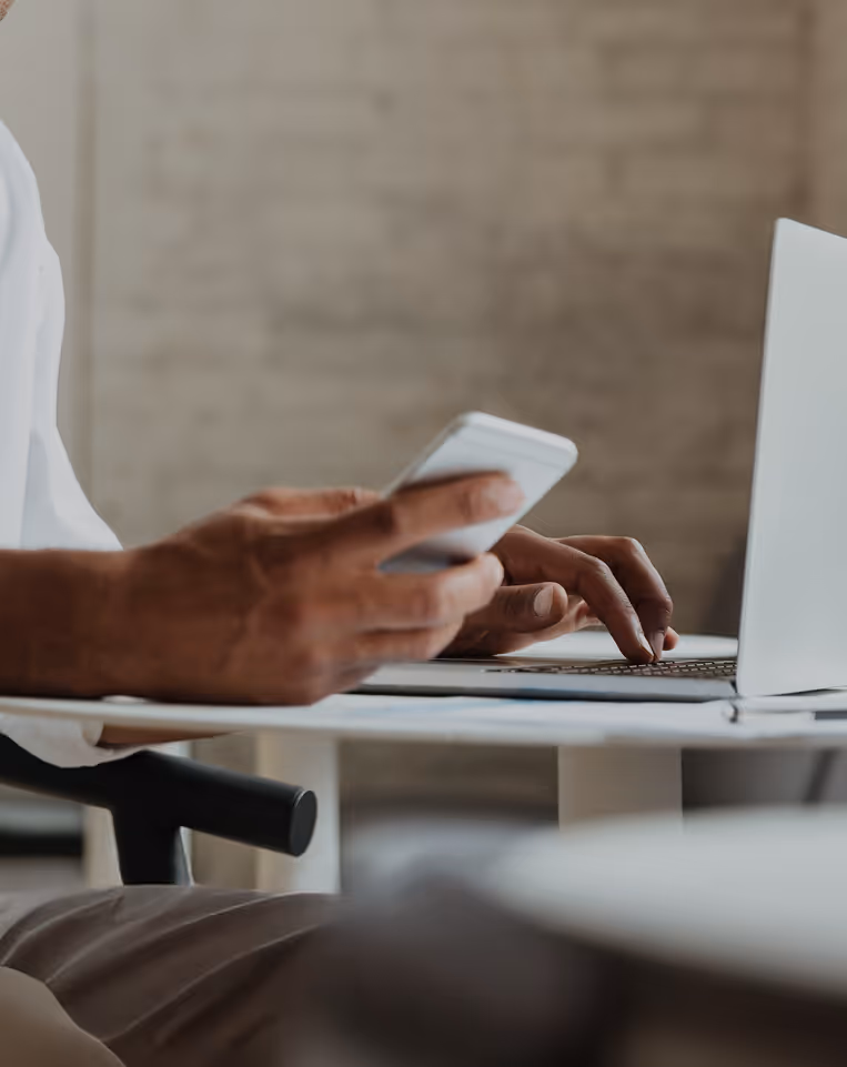 Person using a smartphone while sitting at a desk with a laptop.